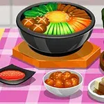 Tazón de sopa con verduras y champiñones, acompañado de té verde, tofu, kimchi y albóndigas sobre un mantel a cuadros rosas