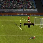 Jugador en campo de fútbol realizando un tiro mientras un árbitro y jugadores observan, con un portero sobre un trampolín frente a la portería