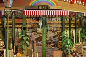 Cafetería con fachada de cristal, rayas rojas y blancas, decorada con plantas verdes y un arco iris en la parte superior