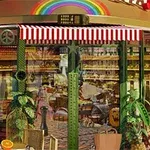 Cafetería con fachada de cristal, rayas rojas y blancas, decorada con plantas verdes y un arco iris en la parte superior